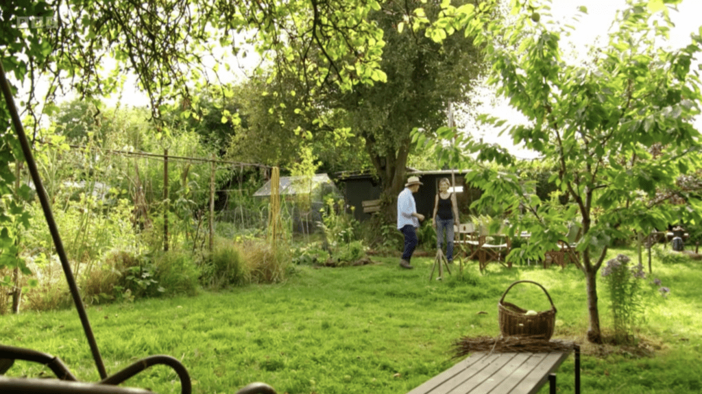 Male and female figure stand chatting in a relaxed pose in the background of a wider shot showing an allotment. Lots of fruit trees, climb frames for beans and different arrangements of seating.