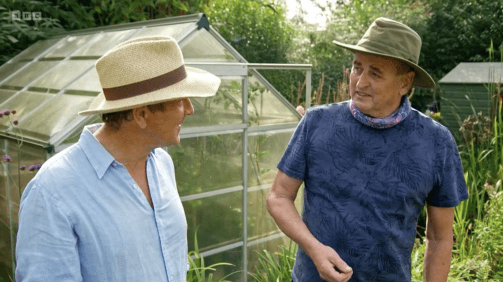 Two men talking while stood in front of a greenhouse. A hedge, a planted border and a wooden shed can be seen in the background.