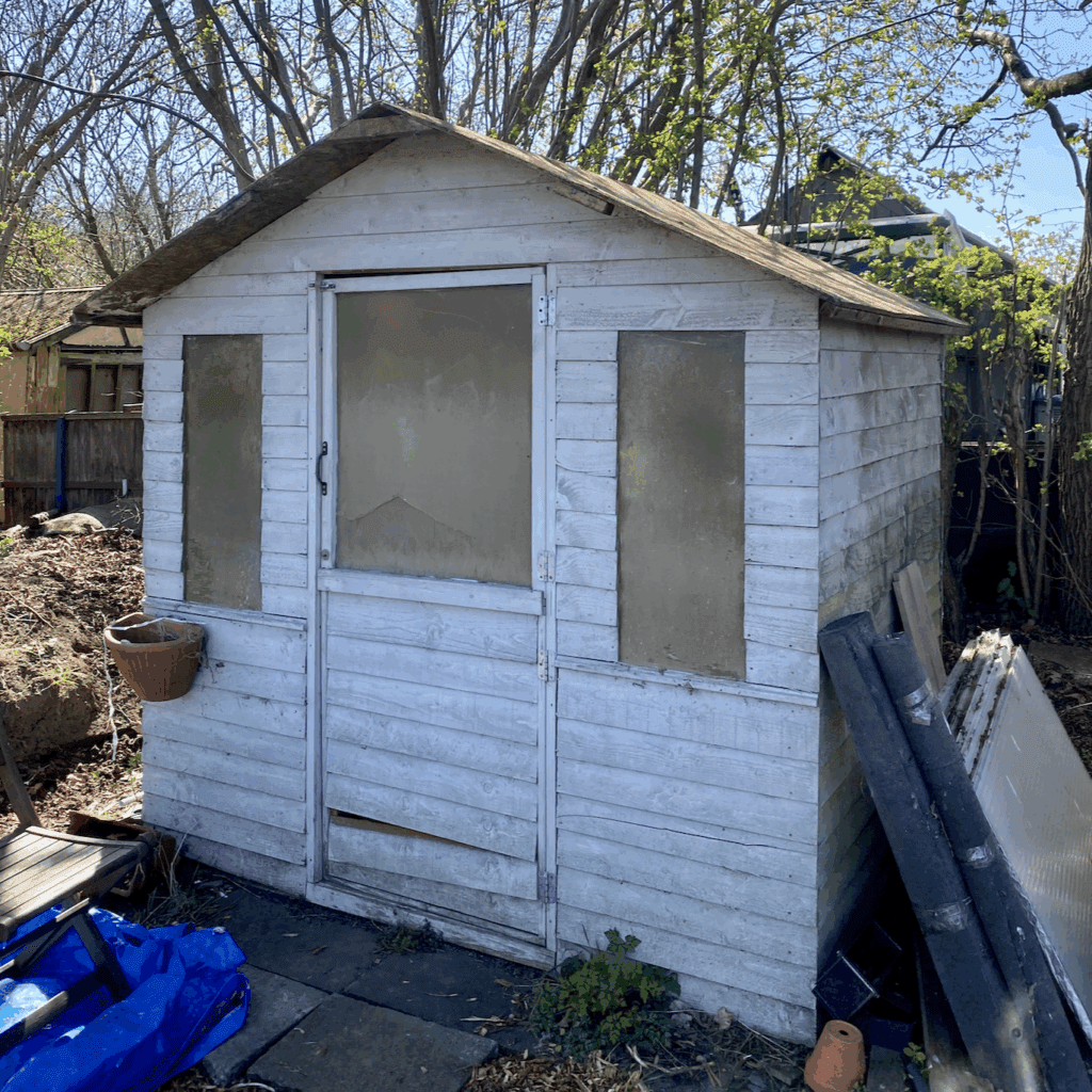 A dilapidated 6ft wide by 4ft deep shed. There is a window either side of the central door. The door also has a window. Boards are coming loose on the door and sides, and the roof is bare OSB without felt.